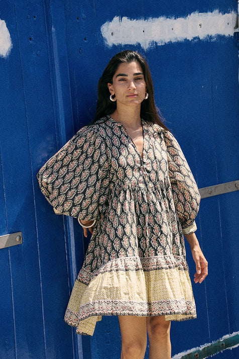 Woman against a blue painted wall wearing the Daughters of India Bhoomi Mini Dress in Onyx, showcasing the dark block print pattern and tiered skirt detail
