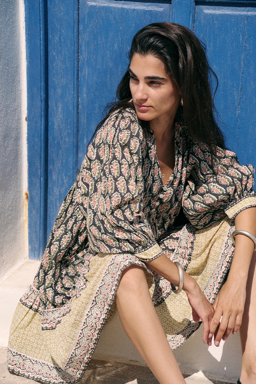 Woman sitting against a blue painted door wearing the Daughters of India Bhoomi Mini Dress in Onyx, showcasing the relaxed boho silhouette and tiered skirt