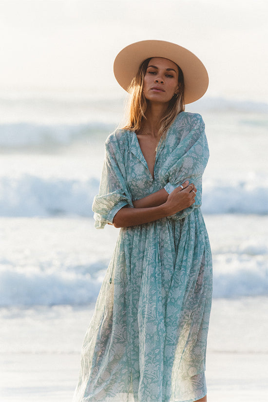 Woman wearing Daughters of India Kyra Midi Dress in Jade with a wide-brim sun hat, standing on a beach with ocean waves in the background