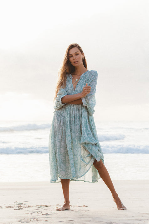 Woman wearing Daughters of India Kyra Midi Dress in Jade, a floral blue-green chiffon dress, standing on a beach