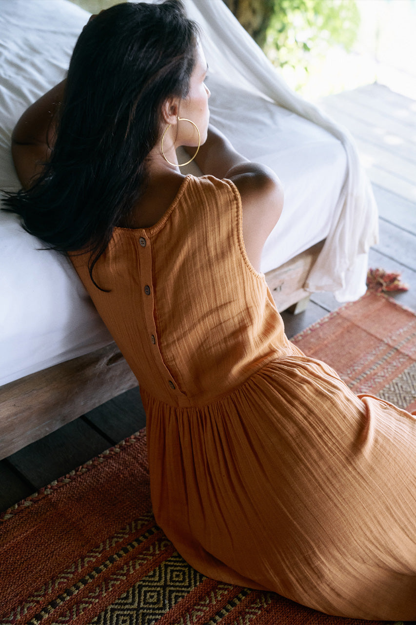 Nadi Dress Amber — back view seated by daybed, amber double gauze cotton midi dress showing back button detail and gathered skirt, tropical resort setting | Daughters of India