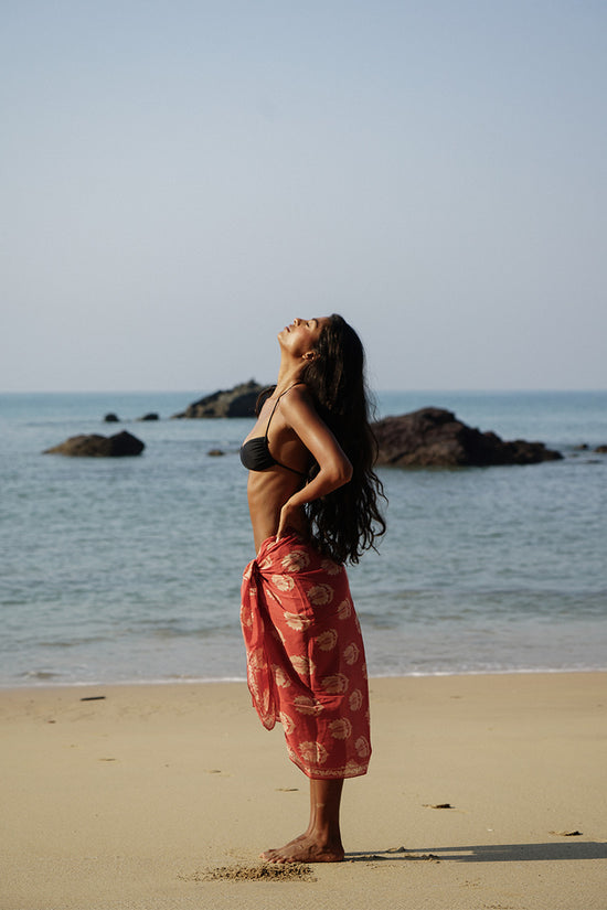 Ria Sarong Ruby Red — side profile on beach looking up at sky, ruby red hand block print cotton sarong tied at waist, black bikini top, ocean and rocks background India | Daughters of India