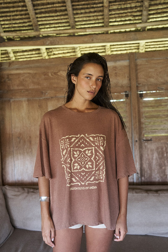 Woman standing in a rustic setting wearing the Daughters of India Shanti Tee in Hazel, an oversized brown block print t-shirt with bangle bracelet